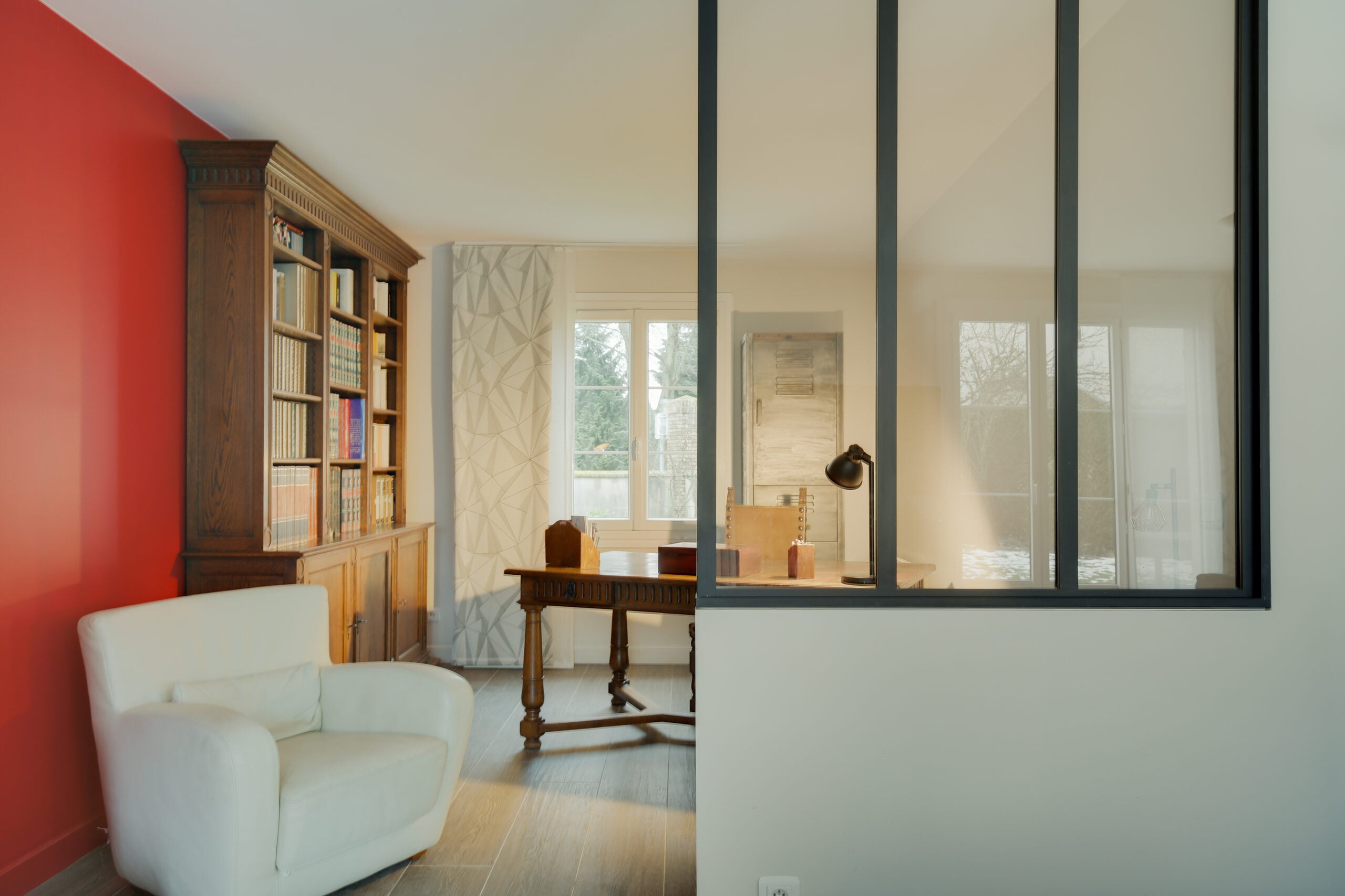 Salon avec mur d'accent rouge, bibliothèque en bois sculpté et verrière noire donnant sur un bureau — réalisation Dec Home