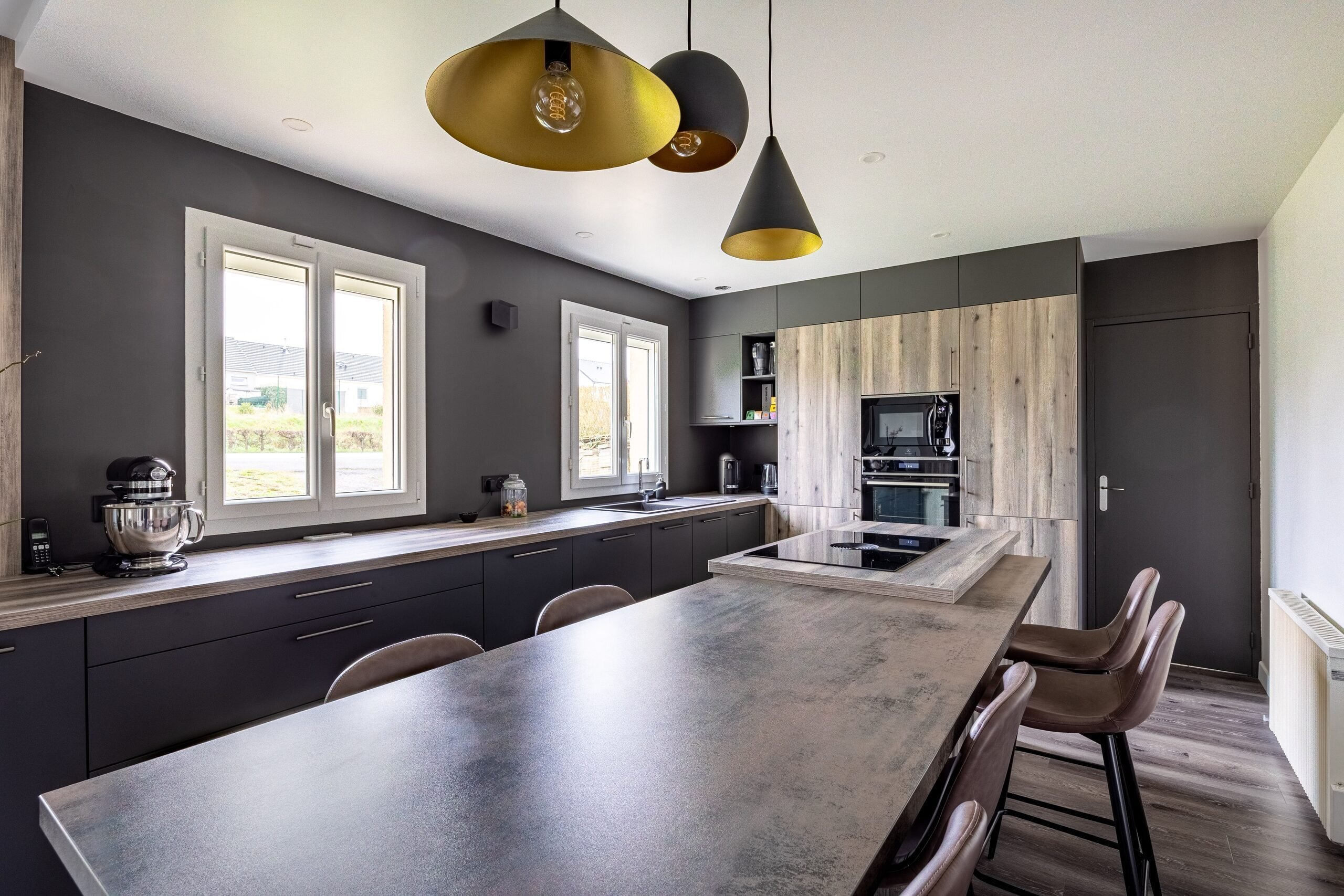 Grande cuisine ouverte avec armoires anthracite, plan de travail béton, colonne en bois et suspension dorée
